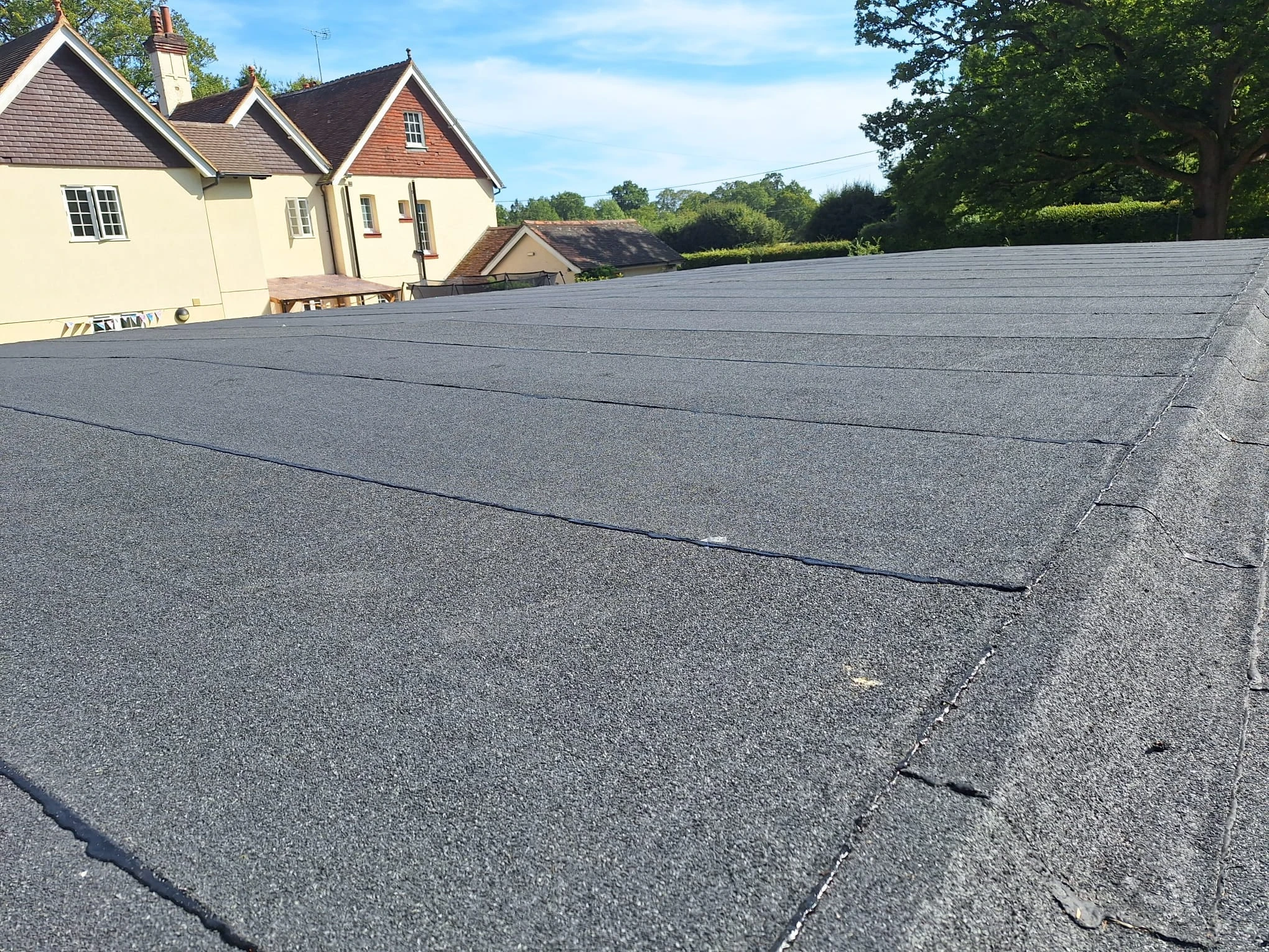 A flat roof covered with neatly arranged dark grey roofing felt panels, with a beige house and green trees in the background under a blue sky.